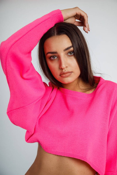 Portrait of young beauteous woman wearing short pink sweatshirt, white trousers, standing on white background, putting hands on head, showing bare belly, posing. Beauty, fashion. Vertical, studio. - Foto, Bild