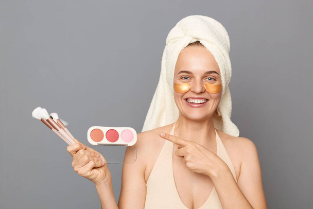 Beauty promotion. Makeup products. Happy woman model with patches under eyes wrapped in towel pointing at powder and brushes advertising isolated over gray background - Foto, Bild