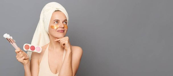 Dreamy pensive woman with patches under eyes wrapped in towel holding powder and brush thinking about today look and makeup isolated over gray background. - Foto, imagen
