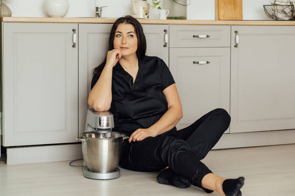 Una mujer cocinera se sienta en el suelo y sostiene una batidora profesional a su lado. Batidora de alta calidad para profesionales - Foto, Imagen