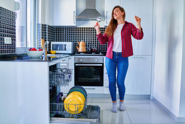 Young joyful happy satisfied smiling cute dancing housewife woman using modern dishwasher for wash dishes and glasses at white home kitchen. Enjoy cleaning chores  - Fotó, kép