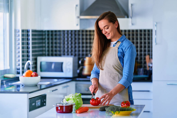 Young happy joyful cute smiling housewife preparing and cutting ripe vegetables on wooden board for fresh vegetarian salad for healthy eating  - Фото, зображення