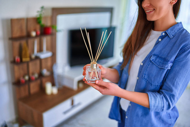 Young happy smiling cute woman enjoying aroma and smell of reed air freshener in the living room at home  - Foto, Bild
