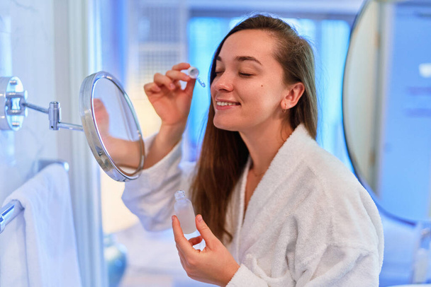 Young beautiful smiling happy female applies a nourishing serum to moisturize skin after shower. Face skin care  - Photo, Image