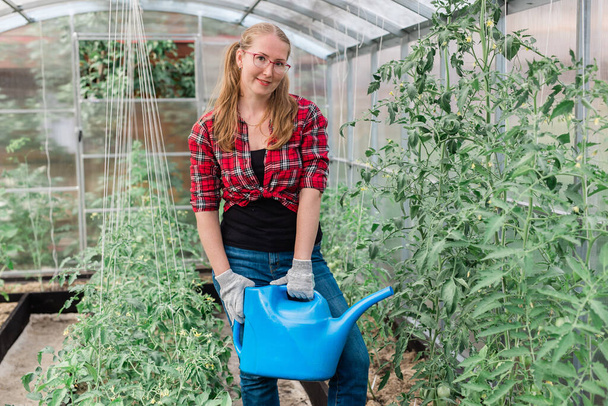 Woman with garden watering can waters plants and green tomatoes, gardening and greenhouse - Foto, afbeelding