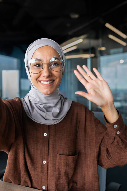 Close-up photo. Vertical photo. A young female blogger in a hijab talks to the camera, holds a phone in her hand. Films a blog, a podcast. He greets the camera with his hand, smiles. - Zdjęcie, obraz