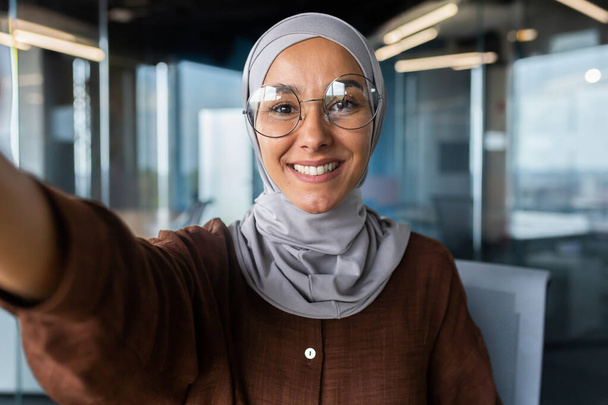 The photo is ugly. Smiling businesswoman in hijab standing in office and talking on video call from phone. Explains, consults, business meeting. - Photo, Image
