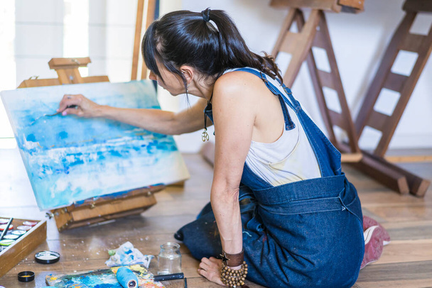 キャンバス上の女性アーティストの絵画青抽象画。彼女のアートスタジオで床で働く創造的な女性. - 写真・画像