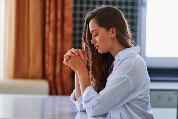 Lonely calm woman believer sits at table with praying hands alone at home and praying to God in silence for support and help. Faith in the best - Zdjęcie, obraz