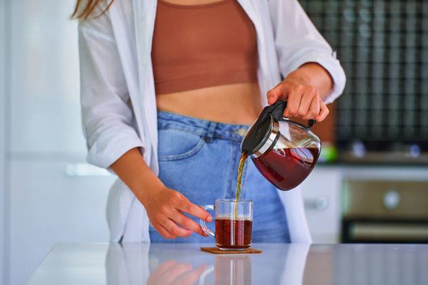 Female pouring black tea from a glass teapot into a cup at home kitchen - Foto, immagini