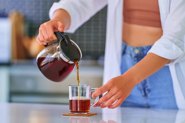Female pouring black tea from a teapot into a glass cup at home kitchen - Photo, image