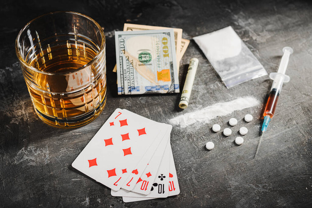Alkoholgetränk im Glas, Karten für Pokerspiele, Spritze mit einer Dosis Drogen, weiße Pillen, Betäubungspulver und US-Dollar-Währung auf dunklem Hintergrund. Konzept von Sucht, Glücksspiel und Missbrauch. - Foto, Bild