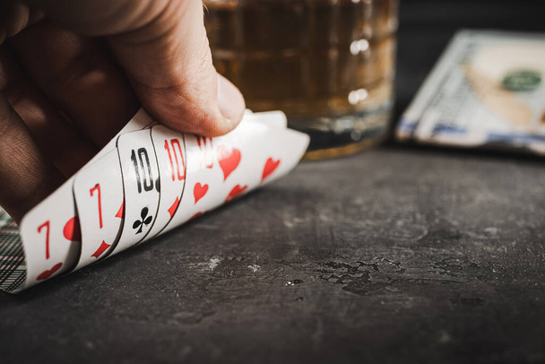 Mies käsi pokerinpelaaja tilalla pelikortteja täyskäsi yhdistelmä, pöydällä dollarin käteisvaluuttaa ja lasi alkoholijuoma. Käsite uhkapeli, riippuvuus ja huonot tavat. - Valokuva, kuva