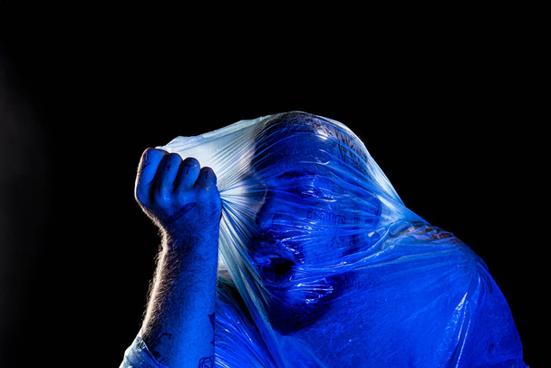 Mężczyzna z plastikową torbą na głowie, uduszony. Studio z niebieskim filtrem. Izolacja na czarnym tle. - Zdjęcie, obraz