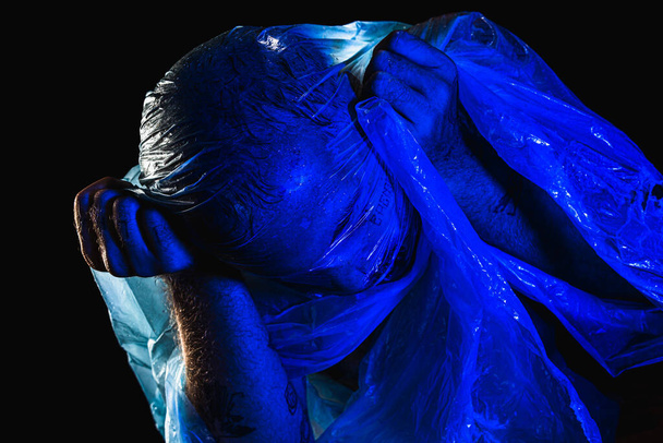 Homme avec un sac en plastique au-dessus de la tête, étouffé. Studio filmé avec filtre bleu. Isolé sur fond noir. - Photo, image