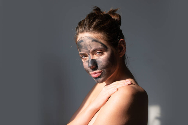 Young woman with naked shoulder and clay mask on face. Beautiful model applying cosmetic cream treatment on face. Facial treatment. Topless woman with black clay mask and bare shoulders - Foto, Imagen