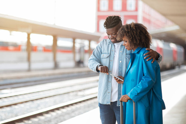 Cónyuges negros felices esperando el tren en la estación de tren, sonriendo hombre y mujer afroamericanos viajando juntos, parados en la plataforma y comprobando la hora de salida en el reloj de pulsera, espacio de copia - Foto, Imagen
