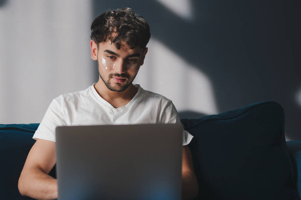 Νεαρός άνδρας που κοιτάζει την οθόνη ενός φορητού υπολογιστή ενώ χαλαρώνει σε έναν άνετο καναπέ στο σπίτι. Εκπαίδευση τεχνολογία Διαδικτύου. Σπιτική άνεση. - Φωτογραφία, εικόνα