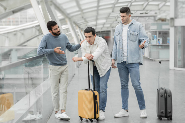 Трое друзей мужчины с дорожным чехлом, имеющие ссору и проблемы во время поездки в ожидании задержки рейса, стоящих в современном аэропорту в помещении. Концепция транспортных вопросов. Full Length Shot - Фото, изображение