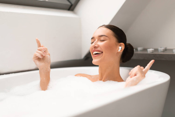 Relaxing Playlist. Relaxed Lady With Earbuds Listening To Music Online Taking Bath With Foam And Singing Favorite Song In Modern Bathroom Indoors, Posing With Eyes Closed. Domestic Relaxation - Photo, image