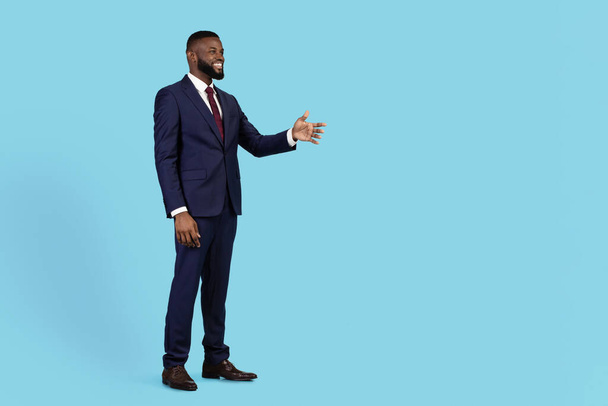 Концепция сотрудничества. Улыбающийся африканский бизнесмен, пожимающий руку за рукопожатие, стоя на фоне синей студии, черный предприниматель в костюме предлагает партнерство, пространство для копирования - Фото, изображение