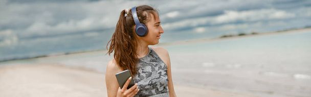 Młoda kobieta w odzieży sportowej słucha muzyki na słuchawkach przy plaży na tle oceanu - Zdjęcie, obraz