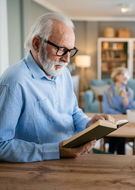 Un uomo maturo anziano caucasico pensionato maschile moderno nonno con capelli corti e barba letto libro a casa indossare occhiali persona reale copia spazio - Foto, immagini
