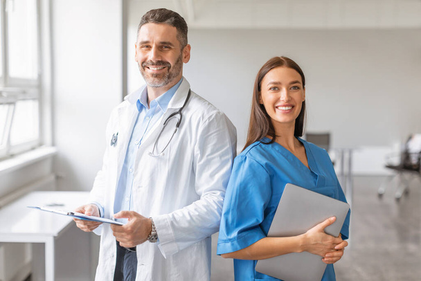 Équipe de deux médecins professionnels travaillant ensemble à la clinique, homme posant avec tablette numérique, femme avec presse-papiers, regardant et souriant à la caméra. Concept de santé et de médecine - Photo, image