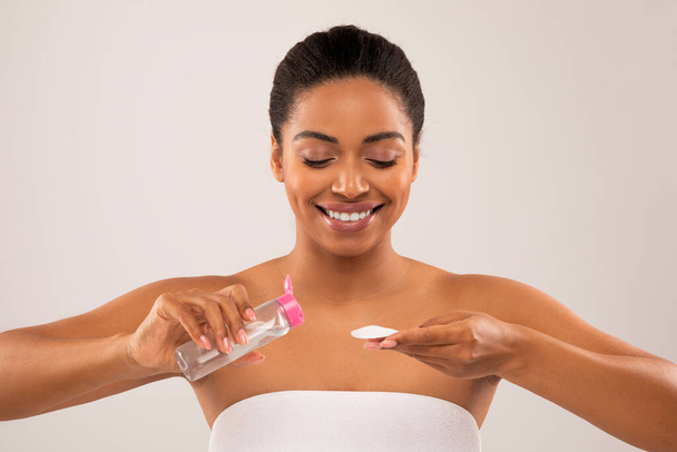 Face care. Attractive cheerful topless millennial black woman using cotton pads and smiling, removing makeup, using toner or micellar water, grey studio background, closeup - Foto, immagini
