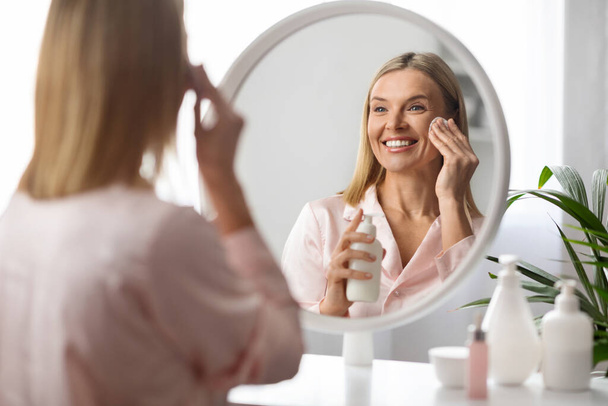 Beautiful Middle Aged Woman Cleansing Skin With Cotton Pad And Makeup Removal Milk, Smiling Attractive Mature Female Doing Daily Beauty Routine While Looking In Mirror At Home, Selective Focus - Foto, immagini