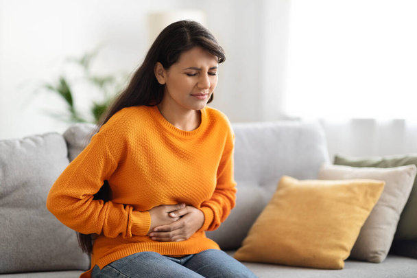Concerned unhappy young indian woman in casual suffering from stomach-ache, sitting on sofa at home. Female holding tummy, undergoing belly pain and discomfort, suffer from menstruation, copy space - Foto, Bild