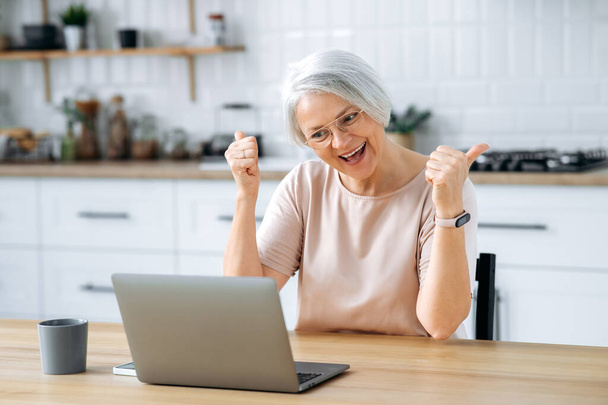 Success, thumb up gesture. Joyful mature caucasian gray haired woman, with glasses, sits in the kitchen at a table with laptop, works remotely, pays bills, manages finances, rejoices at result, smiles - Foto, Bild