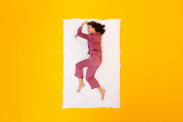 Donna che gode di sonnellino sano che abbraccia cuscino sdraiato su una coperta bianca su sfondo giallo Studio, indossando pigiami. Concetto di routine e ricreazione per dormire. Vista dall'alto - Foto, immagini
