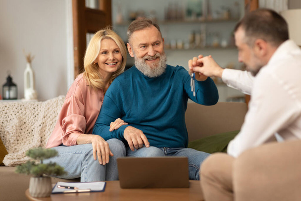 Real Estate Offer. Male Agent Giving Keys From New Home To Cheerful Senior Spouses Buying Own Property Sitting On Couch Indoor. House Purchase And Rent Concept. Selective Focus - Fotografie, Obrázek