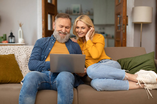 Cónyuges mayores felices usando el ordenador portátil viendo películas en línea sentados en el sofá en casa el fin de semana. El marido y la esposa pasan el tiempo navegando por Internet en la computadora interior. Tecnología y Gadgets - Foto, Imagen
