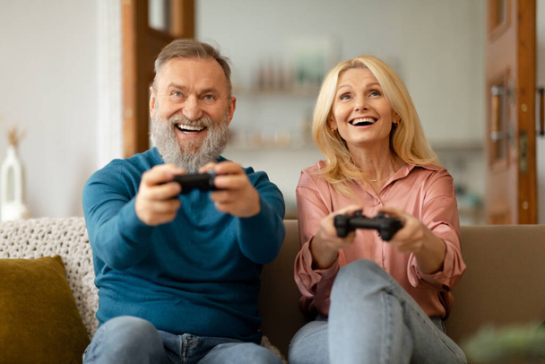 Videojuego. Emocionada pareja mayor jugando videojuegos en línea que sostiene controladores de juegos que se divierten juntos sentados en el sofá en casa el fin de semana. Concepto de Tecnología y Ocio - Foto, Imagen