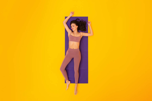 Workout Burnout. Tired Fitness Woman With Eyes Closed Lying On Gymnastic Mat Exhausted After Training Posing Over Yellow Studio Background. Sporty Lifestyle Issues. Full Length Shot - Photo, image