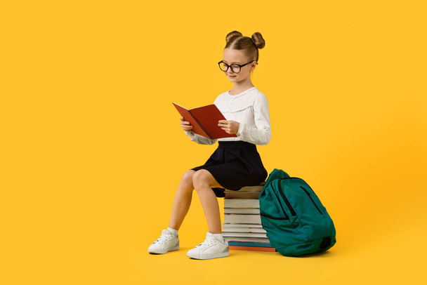 Cute Little Schoolgirl Sitting On Book Stack And Reading Task in Workbook, Beautiful Preteen Female Child Doing School Homework, Enjoying Study, Posing Over Yellow Studio Background, Copy Space - Photo, Image
