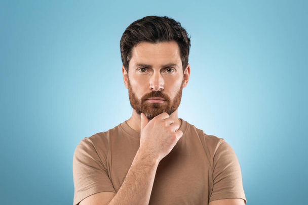 Headshot of attractive focused middle aged man looking at camera and touching unshaven chin, posing on blue background. Skincare cosmetics treatment - Foto, imagen