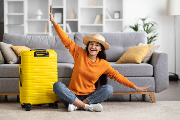 Travel preparation. Cheerful young eastern woman packing suitcase, getting ready for vacation at home, copy space. Female going on tourist trip, happy about abroad adventure, showing airplane - Fotografie, Obrázek