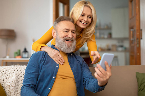Cheerful Senior Couple Using Mobile Phone Texting And Websurfing Sitting On Sofa At Home. Great Application For Your Smartphone. Gadgets And Internet Concept. Selective Focus - Photo, Image