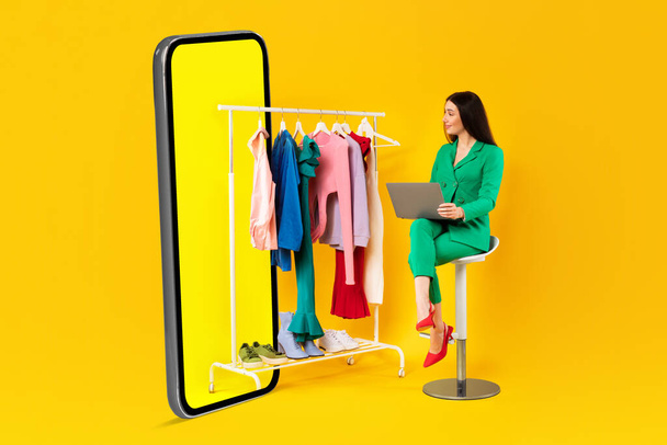 Ενθουσιασμένη γυναίκα σχεδιαστής μόδας με φορητό υπολογιστή κοιτάζοντας τα ρούχα στις ράγες κοντά στο τεράστιο κινητό τηλέφωνο σε κίτρινο φόντο στούντιο. Μόδα, στυλ και σύγχρονη συσκευή - Φωτογραφία, εικόνα