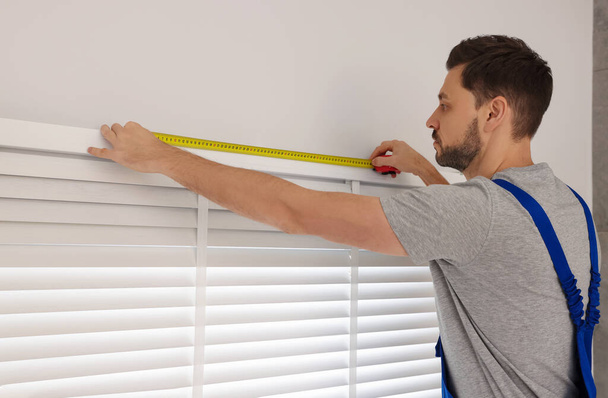 Worker in uniform using measuring tape while installing horizontal window blinds indoors - Foto, Bild