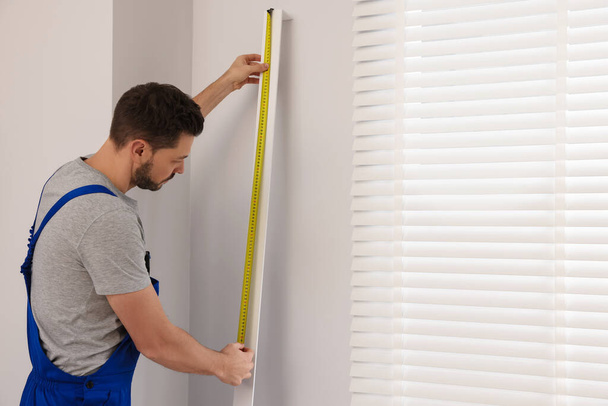 Worker in uniform using measuring tape while installing horizontal window blinds indoors - Foto, afbeelding