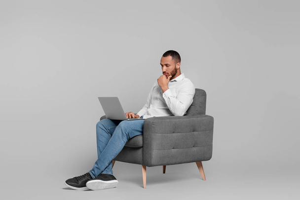 Thoughtful young man with laptop sitting in armchair on grey background - Foto, immagini