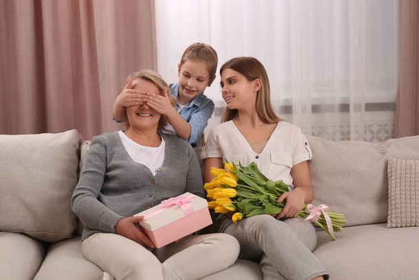 Little girl congratulating her mom and granny with flowers and gift at home. Happy Mother's Day - Foto, Bild