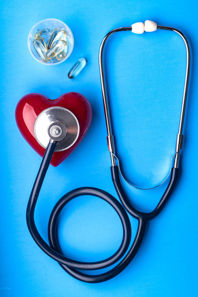 Stetoskop medyczny, pigułki i czerwone serce na niebieskim tle, widok z góry. Miejsce na tekst. - Zdjęcie, obraz