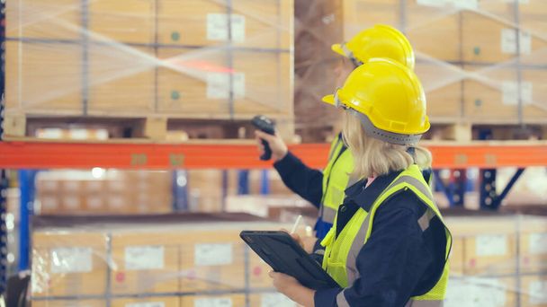 Warehouse workers holding digital tablet checking inventory management packaging boxes. Two staff wearing vest and safety helmet and walking count the box goods on shelves at storehouse - Foto, Bild