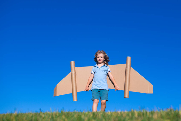Happy child with paper wings against blue sky. Kid with toy jetpack having fun in spring green field outdoor. Freedom carefree and kids imagination dream concept - Photo, Image