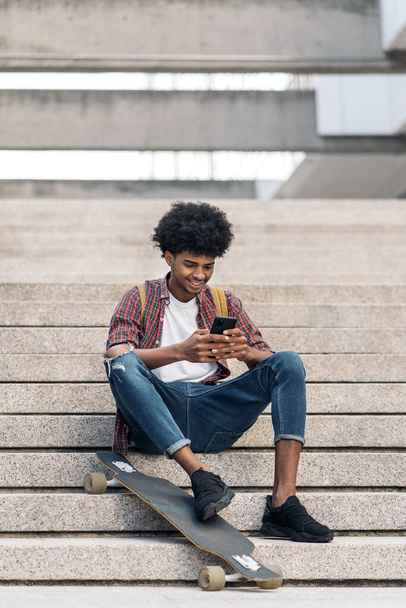 side view, afro boy, using phone, young, male, 20s, afro, cellphone, urban, stairs, sitting, city, outdoors, outside, cool, casual, skater, typing, african american, african, black, ethnic, looking, skate, street, happy, lifestyle, smiling, cheerful, - 写真・画像